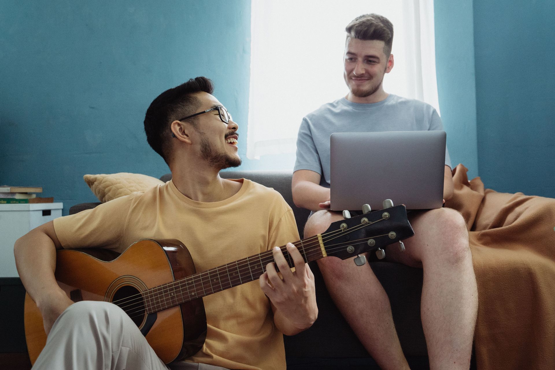 Two friends relax together, one playing guitar and the other using a laptop, creating a joyful atmosphere indoors.