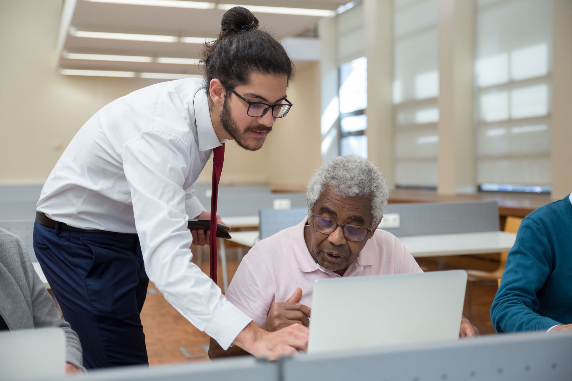 A young man helps a senior adult with laptop use, emphasizing tech education.